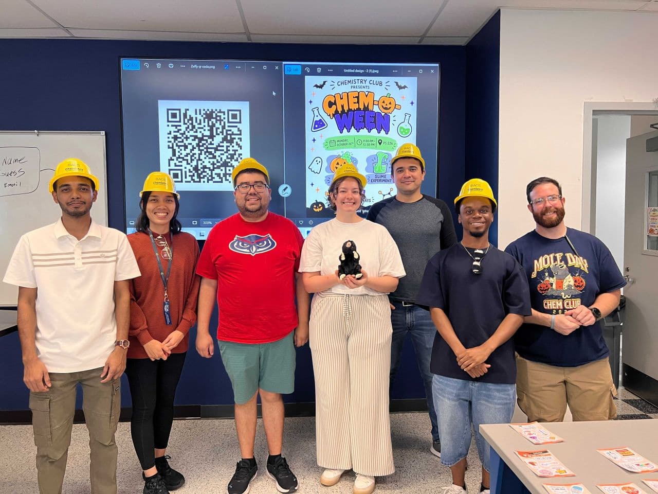 Seven people, several wearing yellow Chemistry hard hats, posing in a classroom in front of a screen advertising the FAU Chemistry Club Chem-o-ween event, one holding a stuffed mole toy